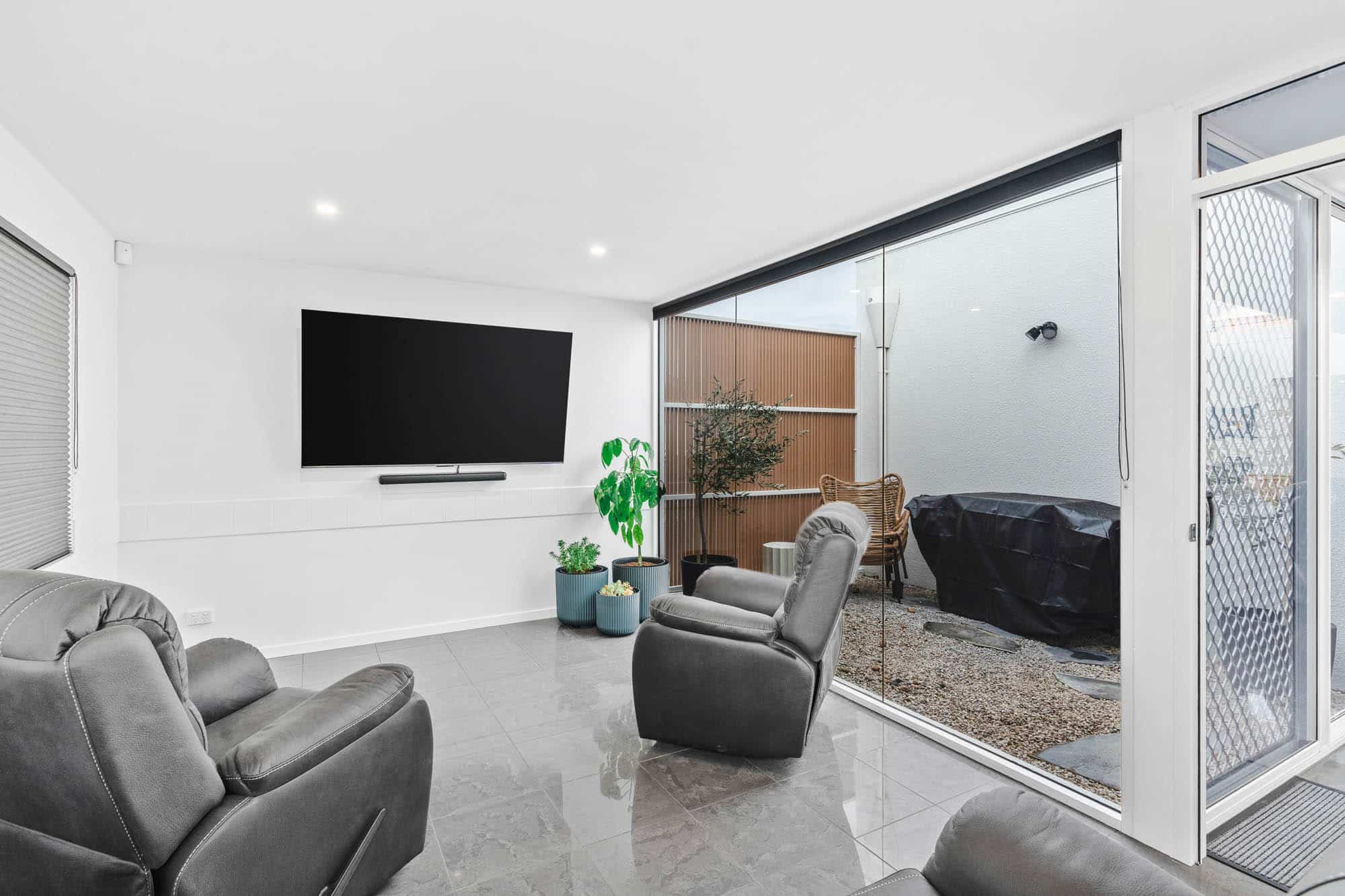 Modern living room with gray recliner chairs, wall-mounted TV, tile flooring, and sliding glass door opening to a small patio with a table, chairs, and covered grill.