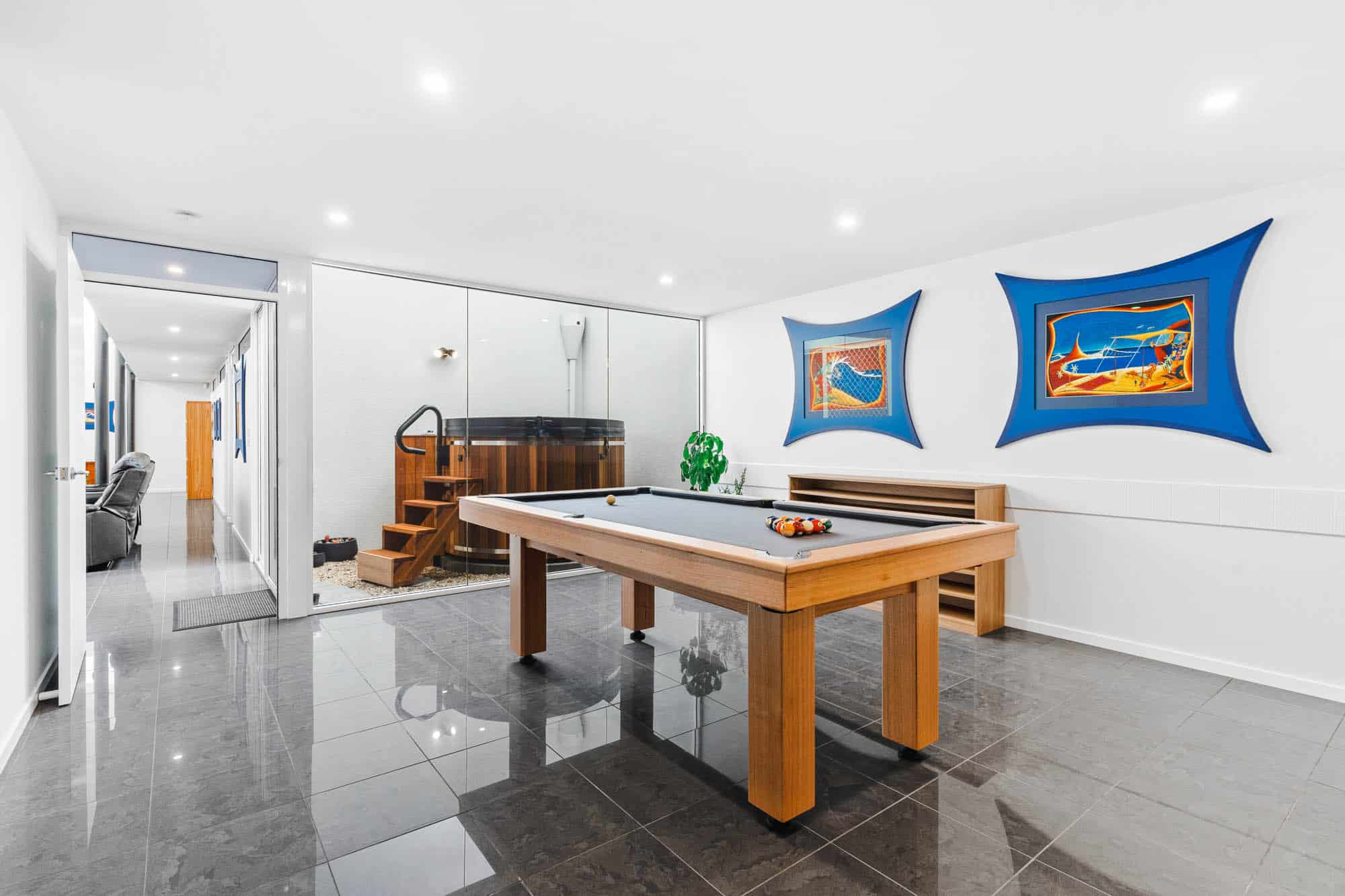 The Prescription Pad-35 Modern game room with a wooden pool table, wall art, glossy tile floors, and a glass wall revealing an outdoor hot tub and staircase.
