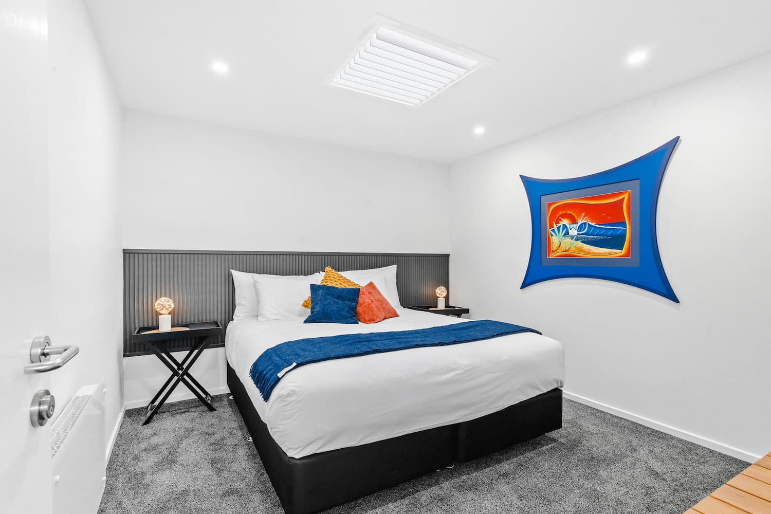 The Prescription Pad5 A modern bedroom with a large bed, white bedding, coloured throw pillows, two small black bedside tables with lamps, grey carpet, white walls, and a colourful framed artwork on the right wall.
