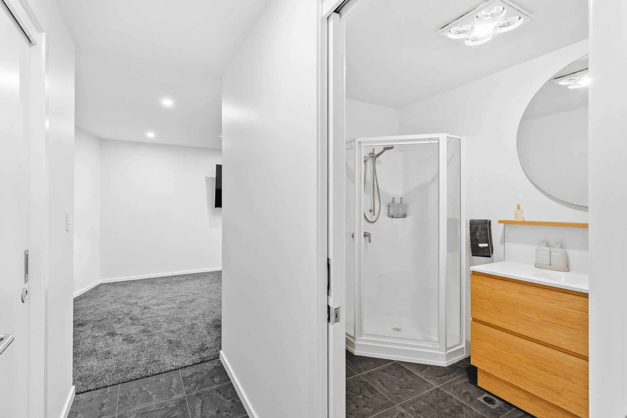 Modern bathroom with a glass shower, wooden vanity, round mirror, and gray tiled floor, adjacent to a carpeted room visible through an open door.