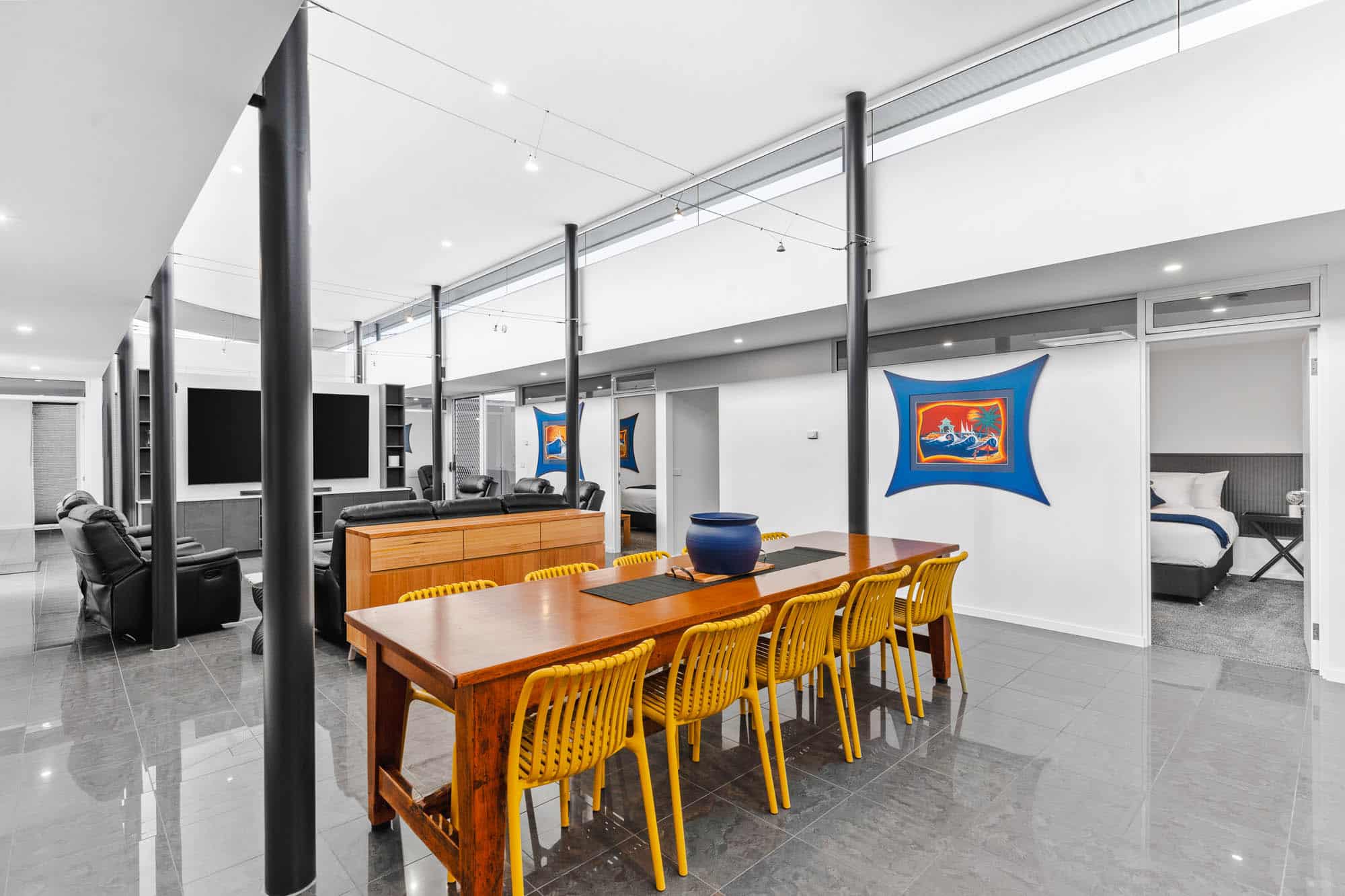 Modern open-plan living and dining area with a wooden table, yellow chairs, black sofas, large TV, and artwork on white walls; bedroom visible in the background.