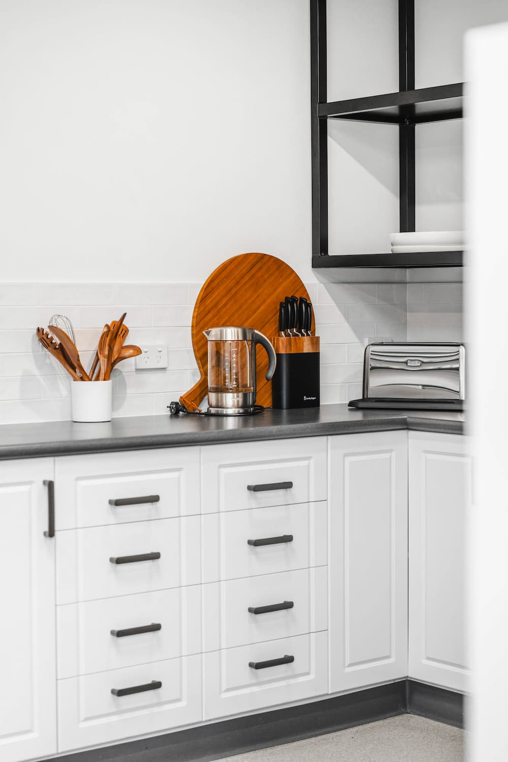 The Prescription Pad2 A modern kitchen corner with white cupboards, black handles, a worktop holding a wooden utensil holder, electric kettle, knife block, wooden chopping board, and a silver toaster. Open black shelves are on the wall above.