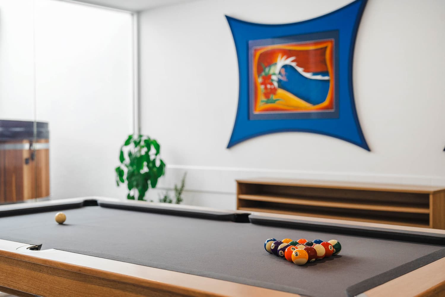 The Prescription Pad4 A pool table with racked billiard balls sits in a modern room. A plant and a wooden shelf are visible in the background, along with a colourful framed abstract artwork on the white wall.