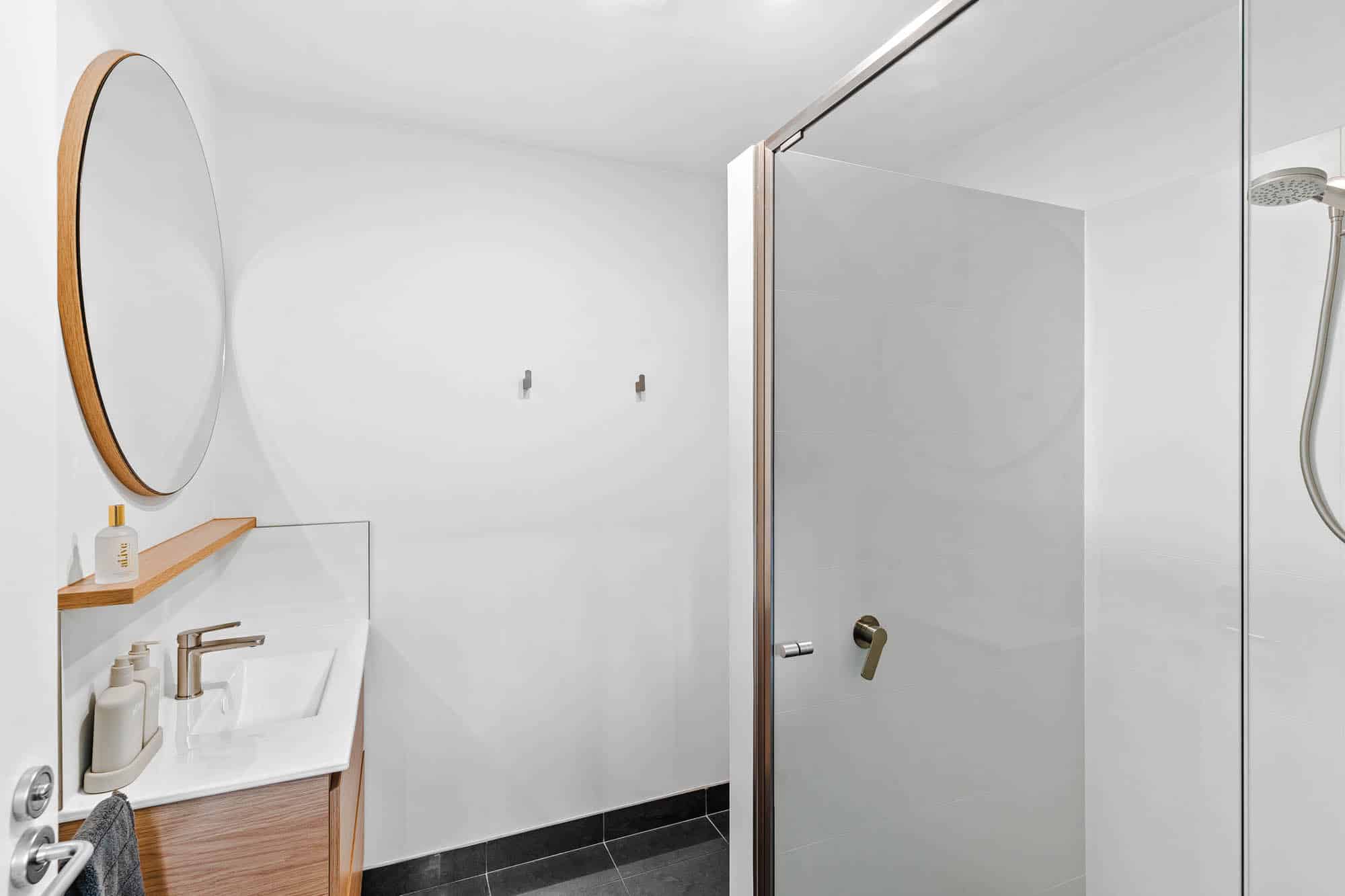 Modern bathroom with a round mirror, small sink with a wooden vanity, wall-mounted fixtures, glass shower enclosure, and minimalistic white and gray design.