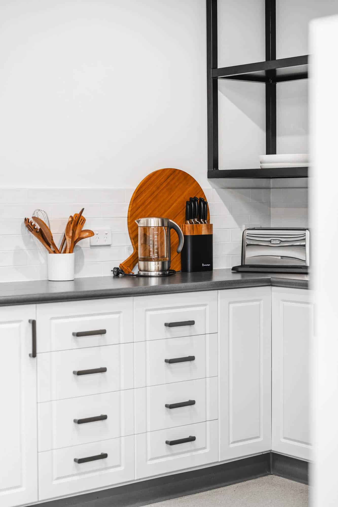 Modern kitchen corner with white cabinets, a kettle, knife block, wooden utensils in a holder, a round cutting board, and a toaster on a gray countertop.