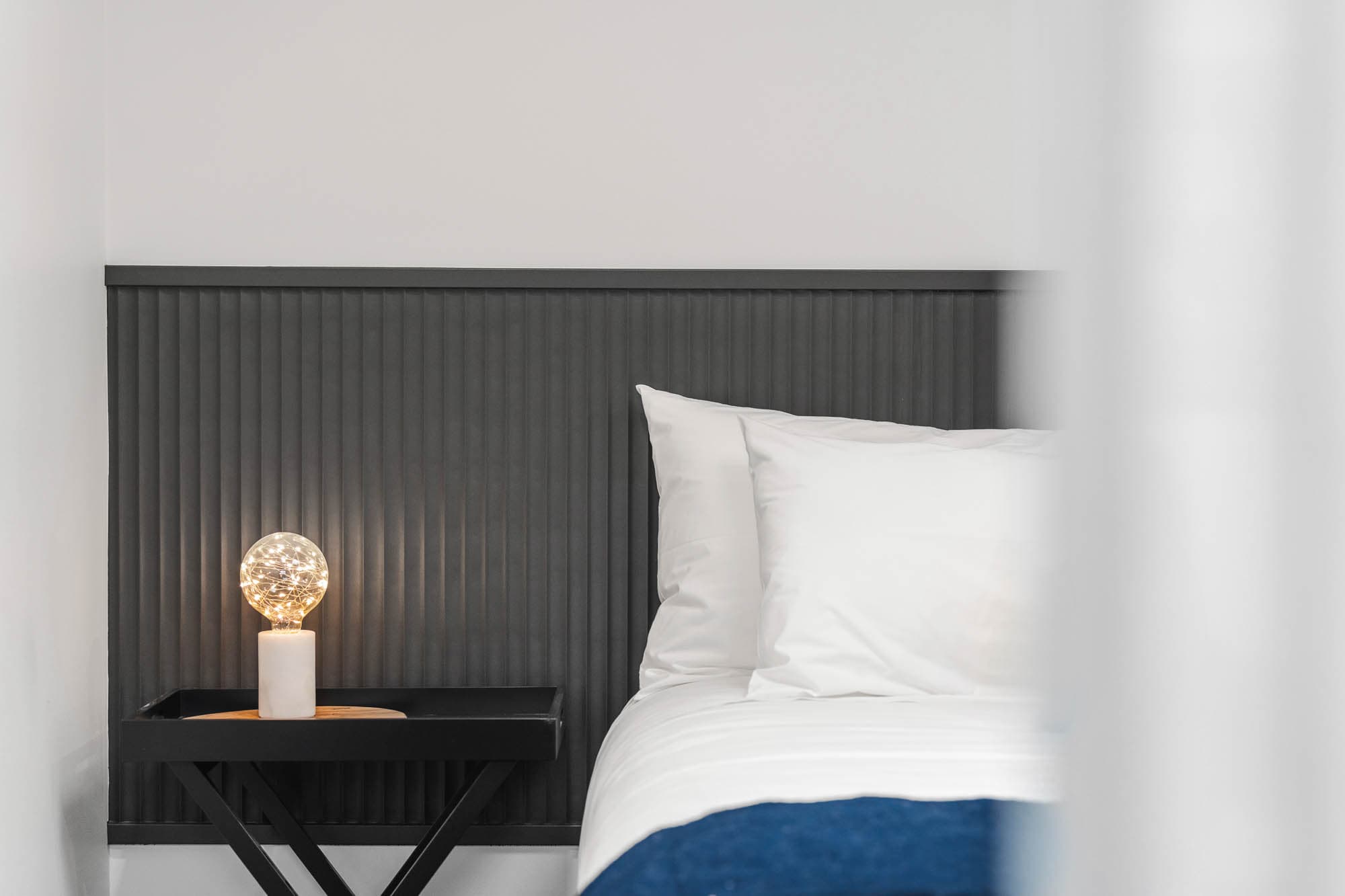 The Prescription Pad Hero 3 A neatly made bed with white linen and a dark grey headboard is shown. A small black bedside table next to the bed holds a lit decorative lamp with a spherical glass shade. The bedding has a blue edge.