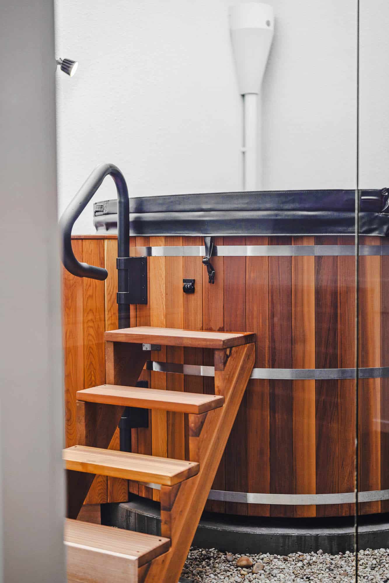A wooden hot tub with metal bands and a black cover, accessed by a small wooden staircase, placed on a gravel surface next to a white wall.