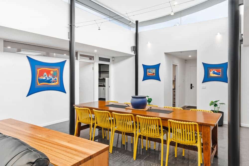 The Prescription Pad1 A modern dining room with a long wooden table, eight yellow chairs, a blue vase centrepiece, white walls, three colourful paintings, black columns, and natural light from skylights above.
